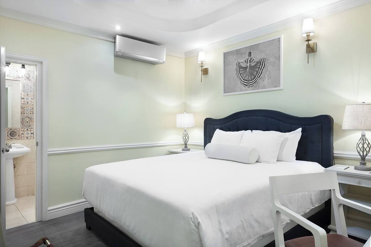 A hotel room with a bed, a chair, a nightstand, a wall-mounted air conditioner, and a decorative wall piece.