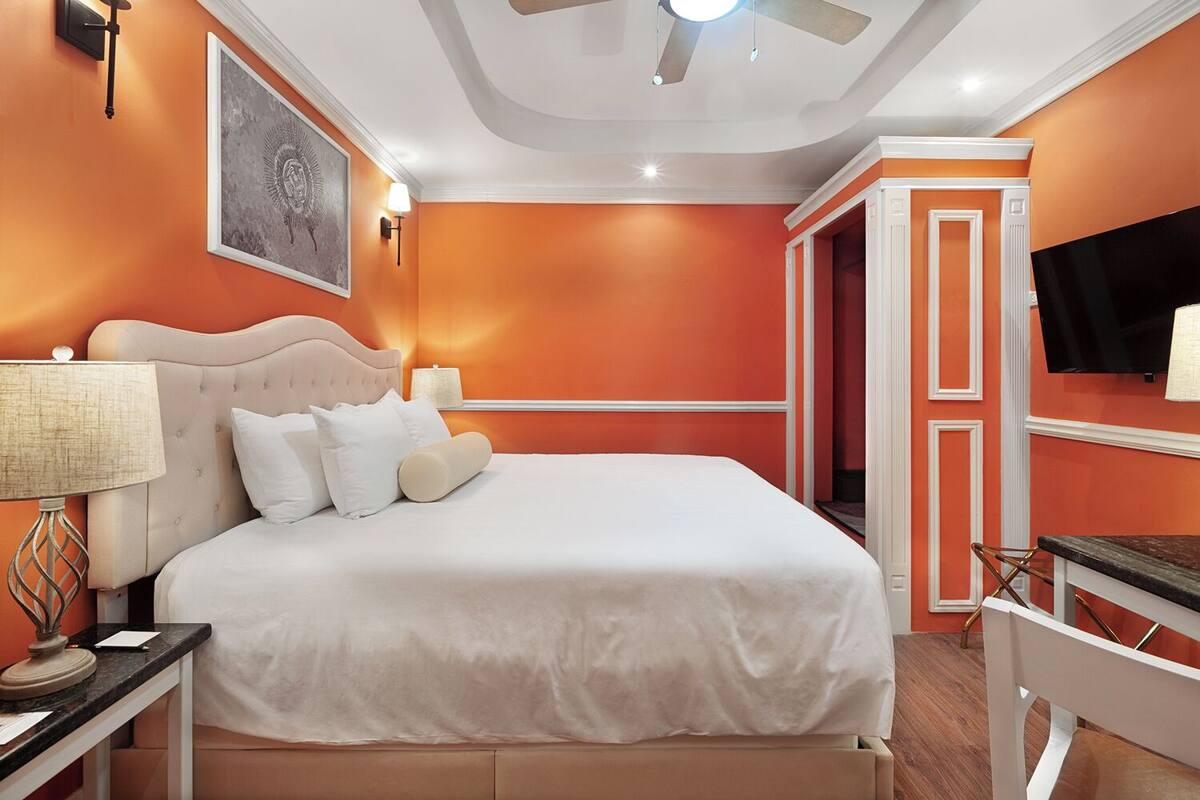 A bedroom with a bed, a nightstand, a lamp, a ceiling fan, a painting, and a TV mounted on the wall.