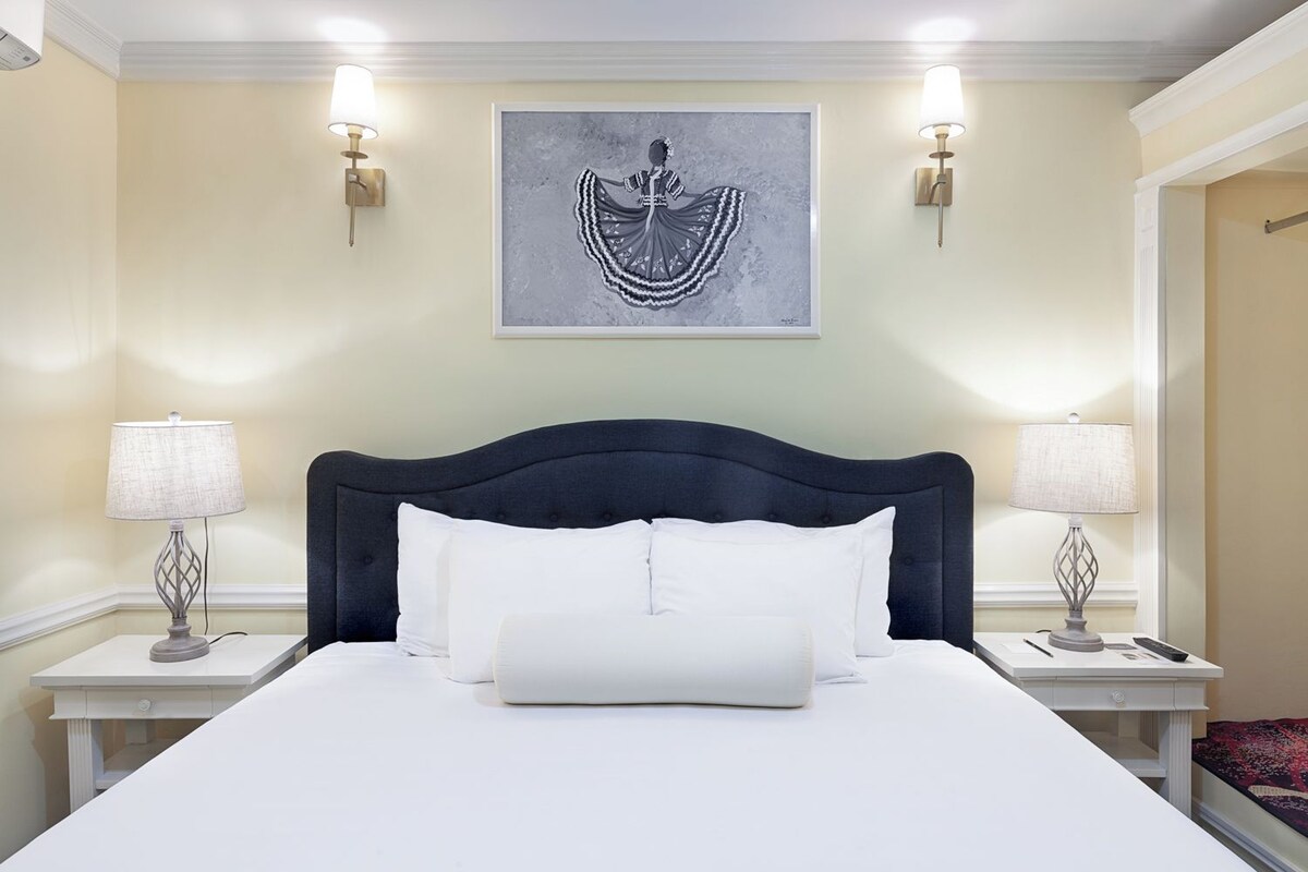A neatly made bed with white linens and a dark headboard, flanked by two bedside tables with lamps, in a room with cream-colored walls and a framed artwork above the bed.