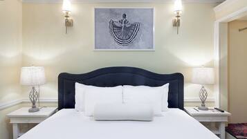 Una cama bien hecha con sábanas blancas y un cabecero oscuro, flanqueada por dos mesitas de noche con lámparas, en una habitación con paredes color crema y una obra de arte enmarcada sobre la cama.