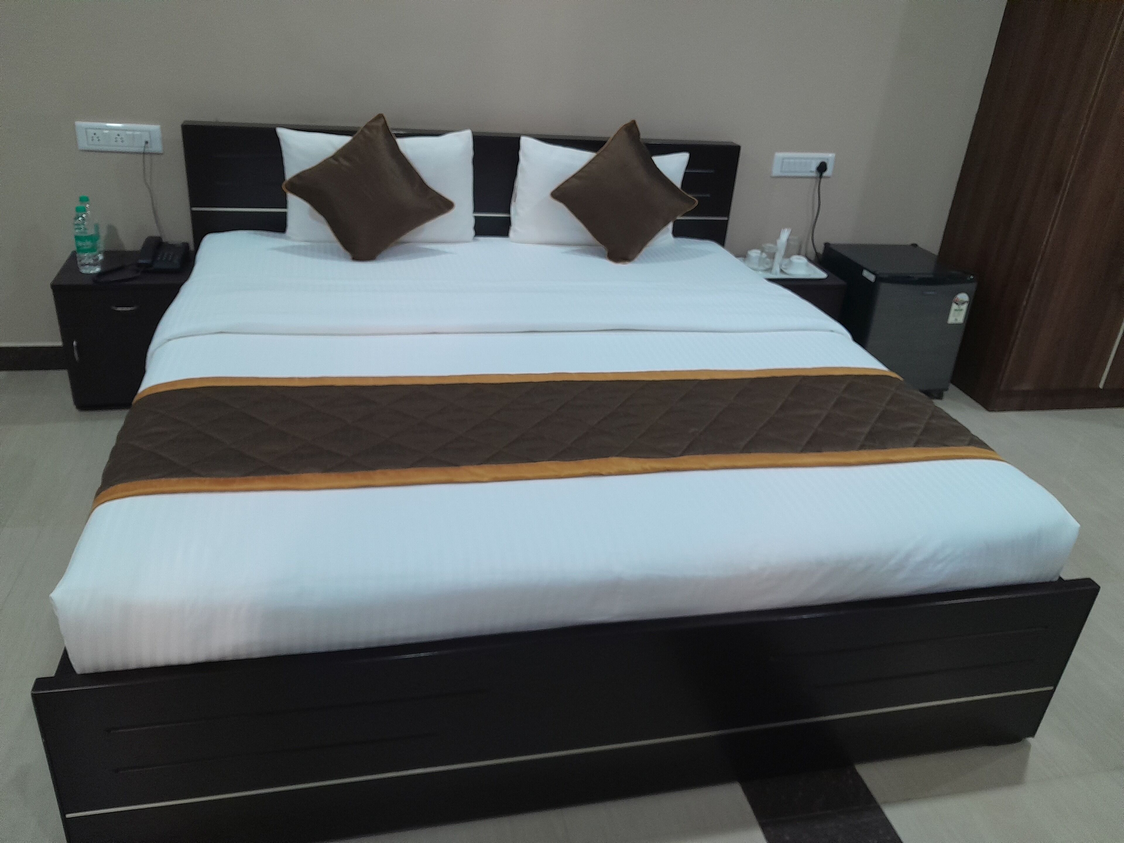 A hotel room with a large bed, a nightstand with a phone and a bottle, a trash bin, and a wooden door.