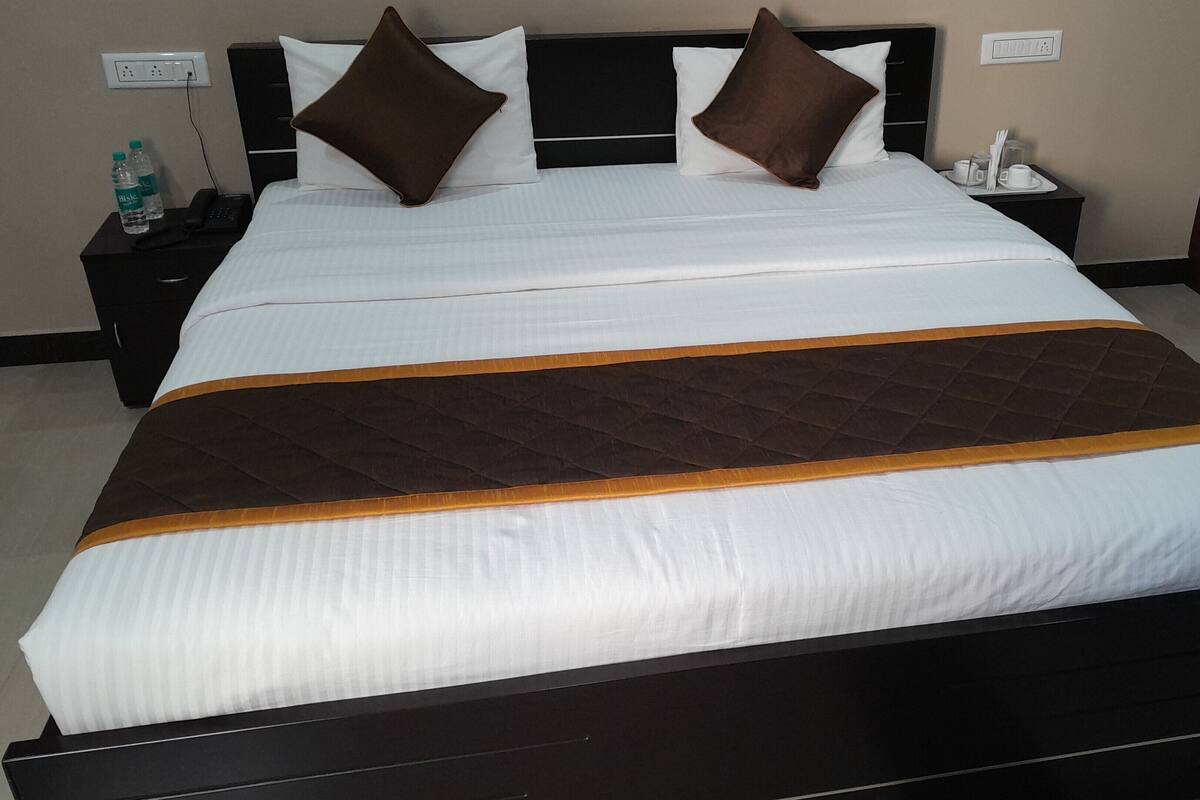 Ein ordentlich bezogenes Bett mit einer gesteppten braunen und weißen Tagesdecke, zwei Kissen, ein Nachttisch mit Telefon und Wasserflaschen sowie eine Steckdose an der Wand.