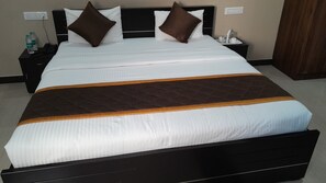Una cama bien hecha con una colcha marrón y blanca acolchada, dos almohadas, una mesita de noche con un teléfono y botellas de agua, y un enchufe en la pared.