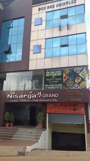 Un edificio de varios pisos, incluyendo una planta baja con restaurante llamado 'Nisarga Grand' y un letrero que dice 'Bee Gee Complex' en la parte superior.