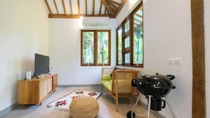 Une pièce avec un plafond en bois, un téléviseur posé sur un meuble en bois, un pouf en fibres tressées, une chaise en rotin et un barbecue.