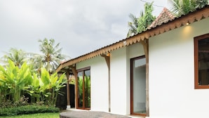 Un bâtiment blanc avec un toit recouvert de tuiles, une table en bois avec quatre tabourets et un petit jardin planté de plantes tropicales.