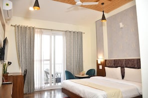 A hotel room with a bed, a desk, a chair, a table, a ceiling fan, and a sliding glass door. - Hotel Le Halcyon Kasau (Solan)