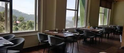 A restaurant with multiple tables and chairs, large windows with a view of greenery and hills.