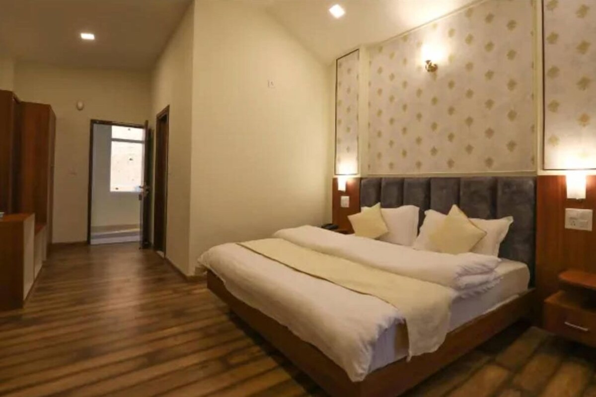 A hotel room with a wooden floor, a large bed with white bedding, a headboard with a patterned wallpaper, and a nightstand with a lamp.