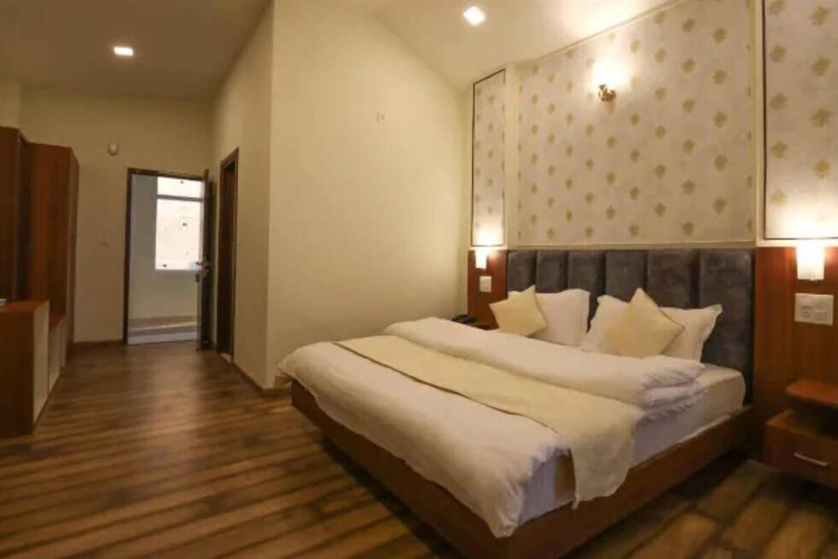 A hotel room with a wooden floor, a large bed with white bedding, a headboard with a patterned wallpaper, and a nightstand with a lamp.