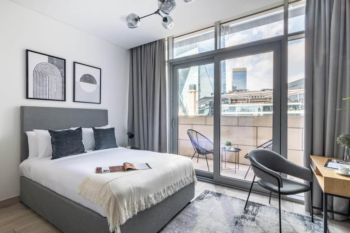 A modern hotel room with a large bed, a desk, and a view of the cityscape through large windows.