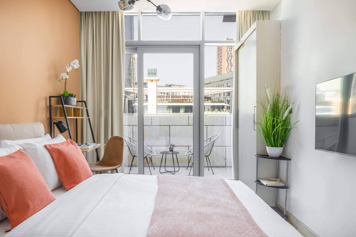 A modern hotel room with a large bed, a TV, a balcony with a view of the city, and a plant on the side table.