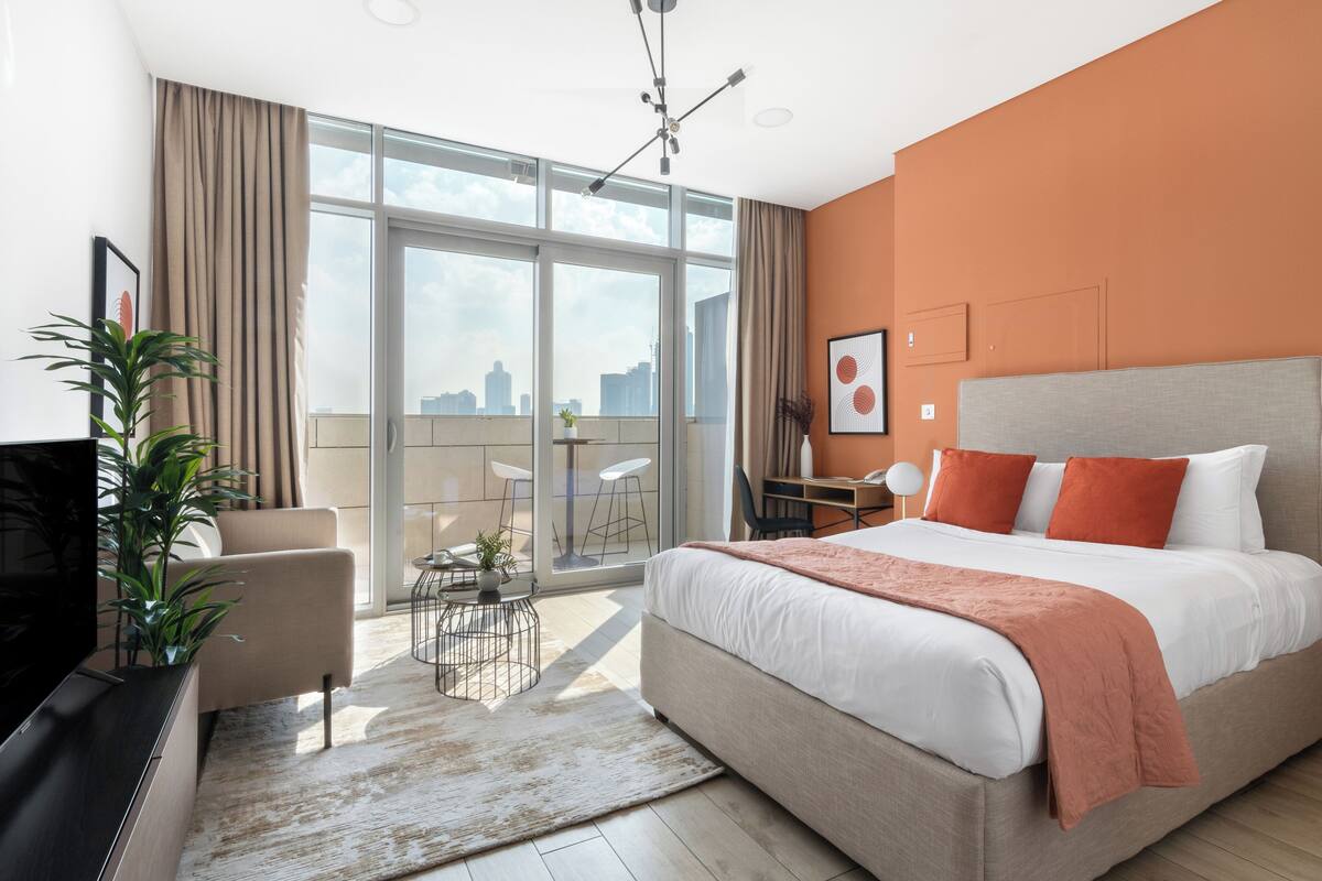 A modern hotel room with a large bed, a desk, a chair, and a balcony with a city view.