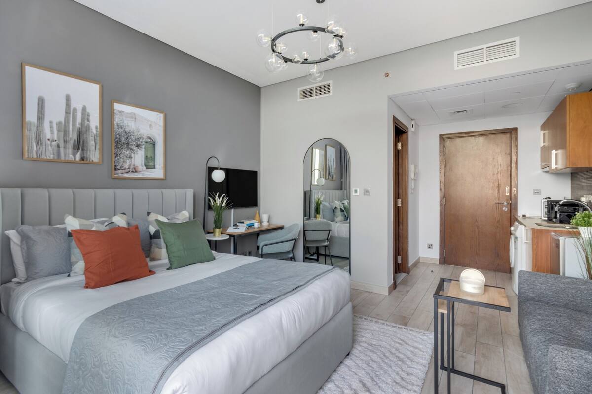 A modern bedroom with a large bed, bedside tables, a desk, and a chair. There is a small kitchen area with cabinets and a microwave.