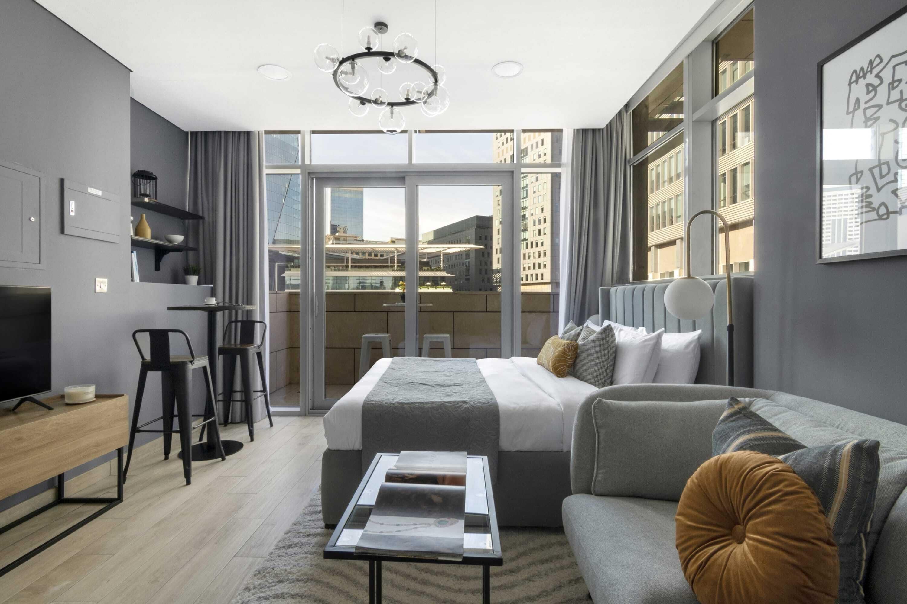 Ein modernes Hotelzimmer mit einem großen Bett, einer Couch, einem Couchtisch und einem Schreibtisch. Große Fenster bieten Blick auf die Stadt.