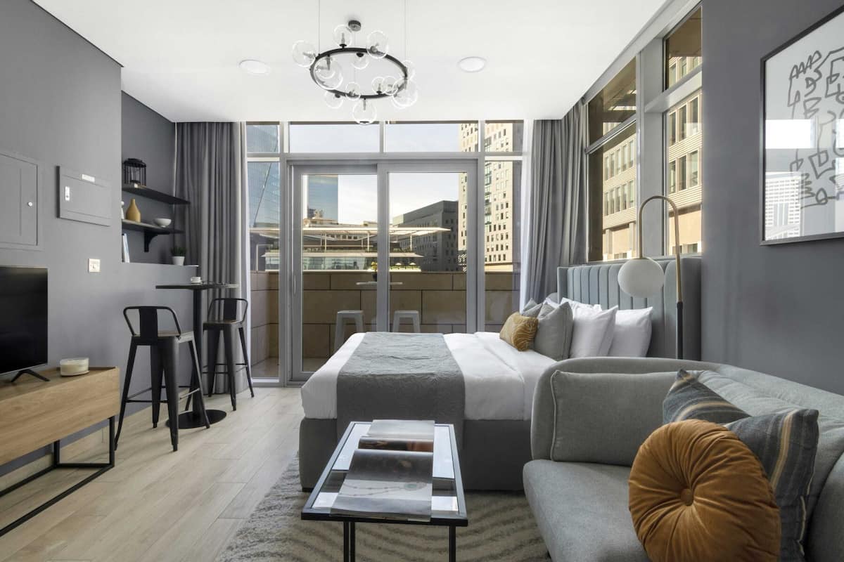 A modern hotel room with a large bed, a sofa, a coffee table, and a desk. There is a view of the city through large windows.