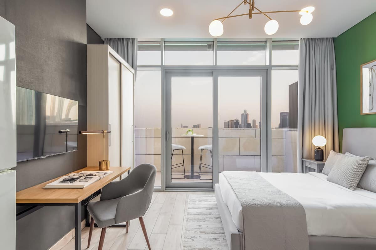 A modern hotel room with a large bed, a desk with a chair, a TV, and a balcony with a city view.