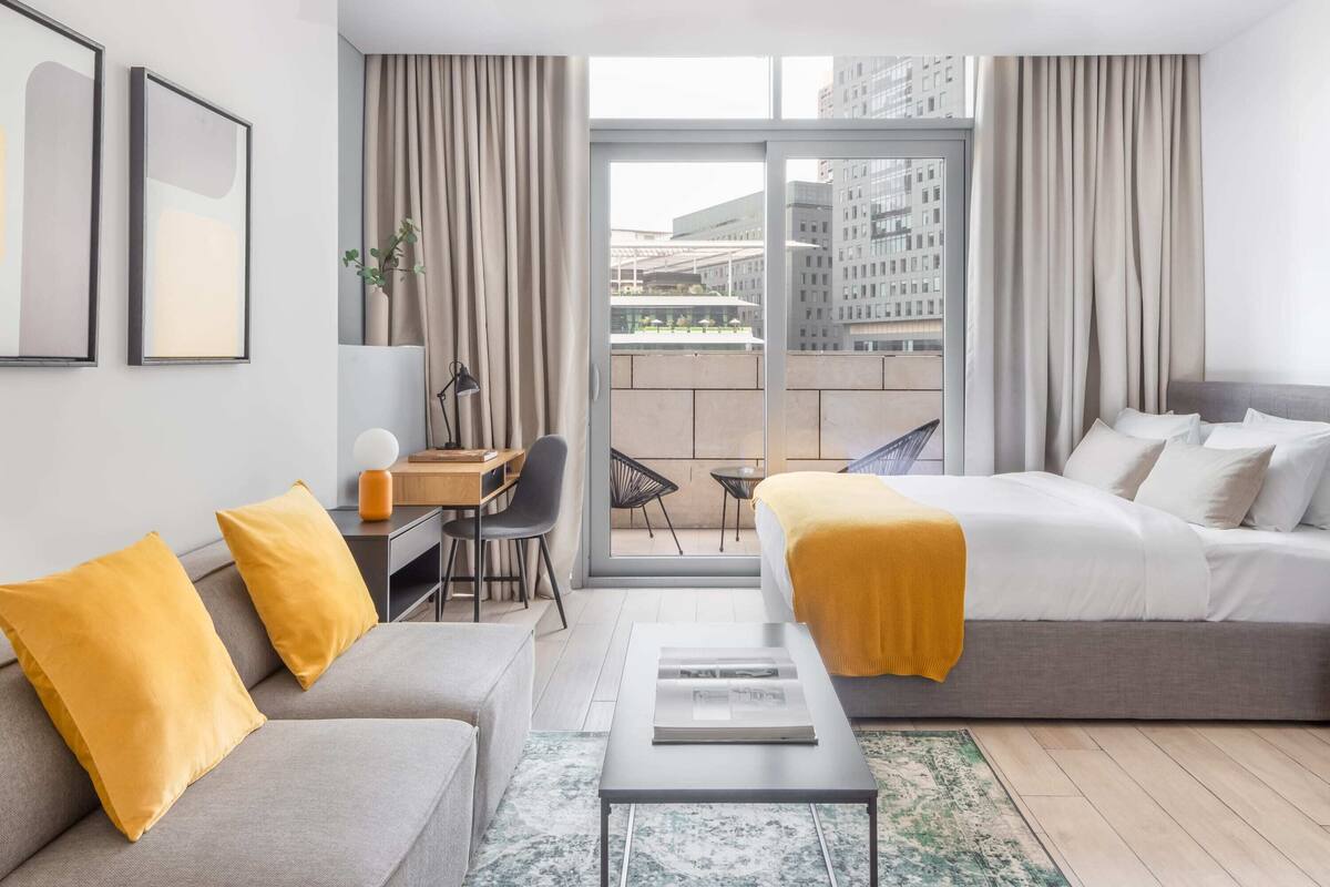 A modern hotel room with a sofa, a bed, a desk, and a view of the city through large windows.