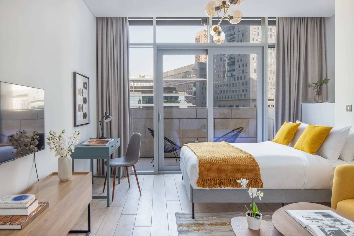 A modern hotel room with a bed, a desk, a chair, a TV, and a view of the city through large windows.
