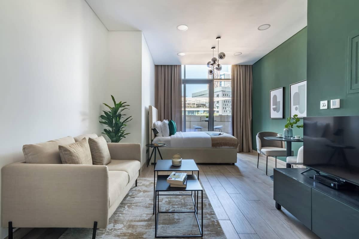 A modern living room with a sofa, coffee table, and TV stand. A bed is visible in the background with a city view.