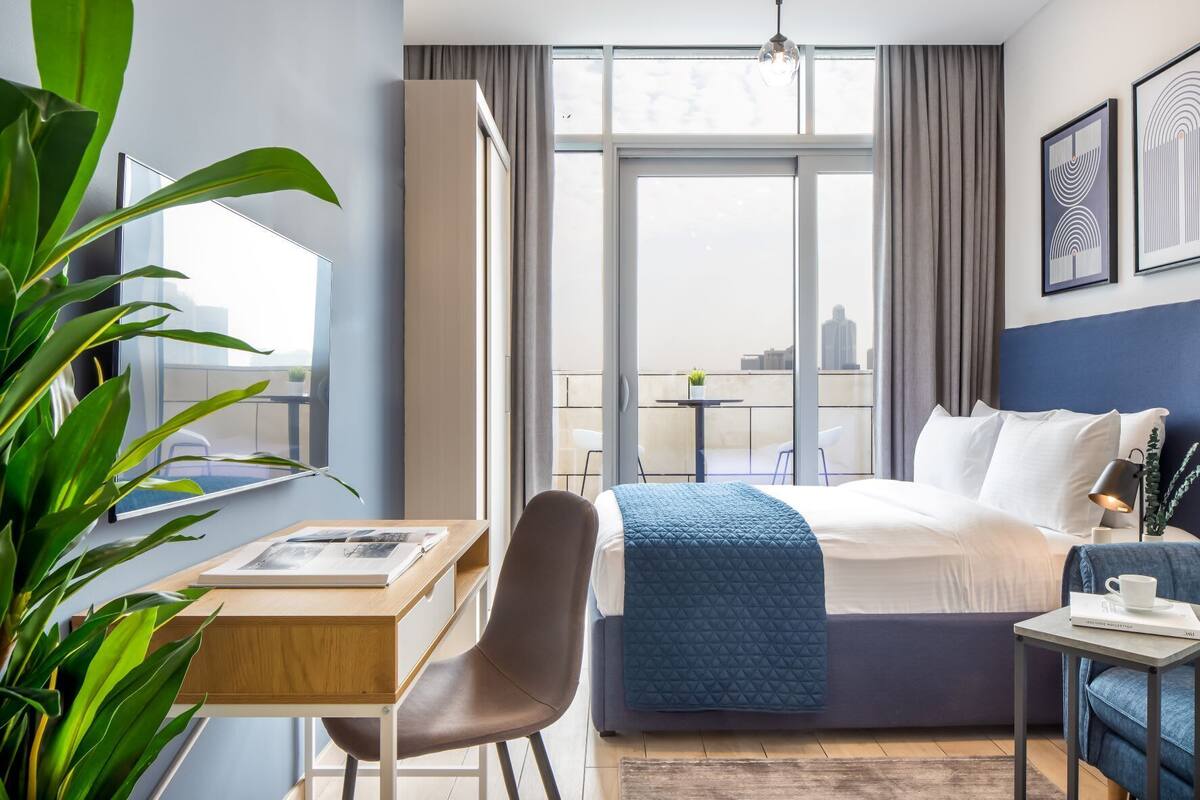 A hotel room with a bed, a desk, a chair, a small table, a potted plant, and a balcony with a view of the city.