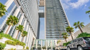 A tall, modern hotel with a glass facade, palm trees, and a parking area. - Burj Daman (Dubai)