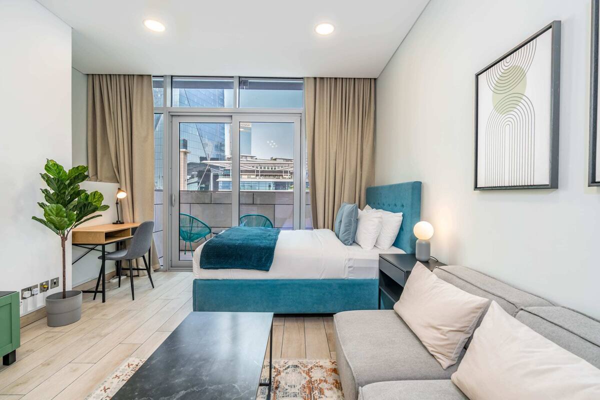 A modern hotel room with a bed, a desk, a chair, a sofa, and a large window with curtains.