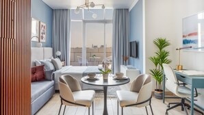 A modern hotel room with a bed, a dining table with chairs, a desk with a chair, a potted plant, and a large window with curtains. - Burj Daman (Dubai)