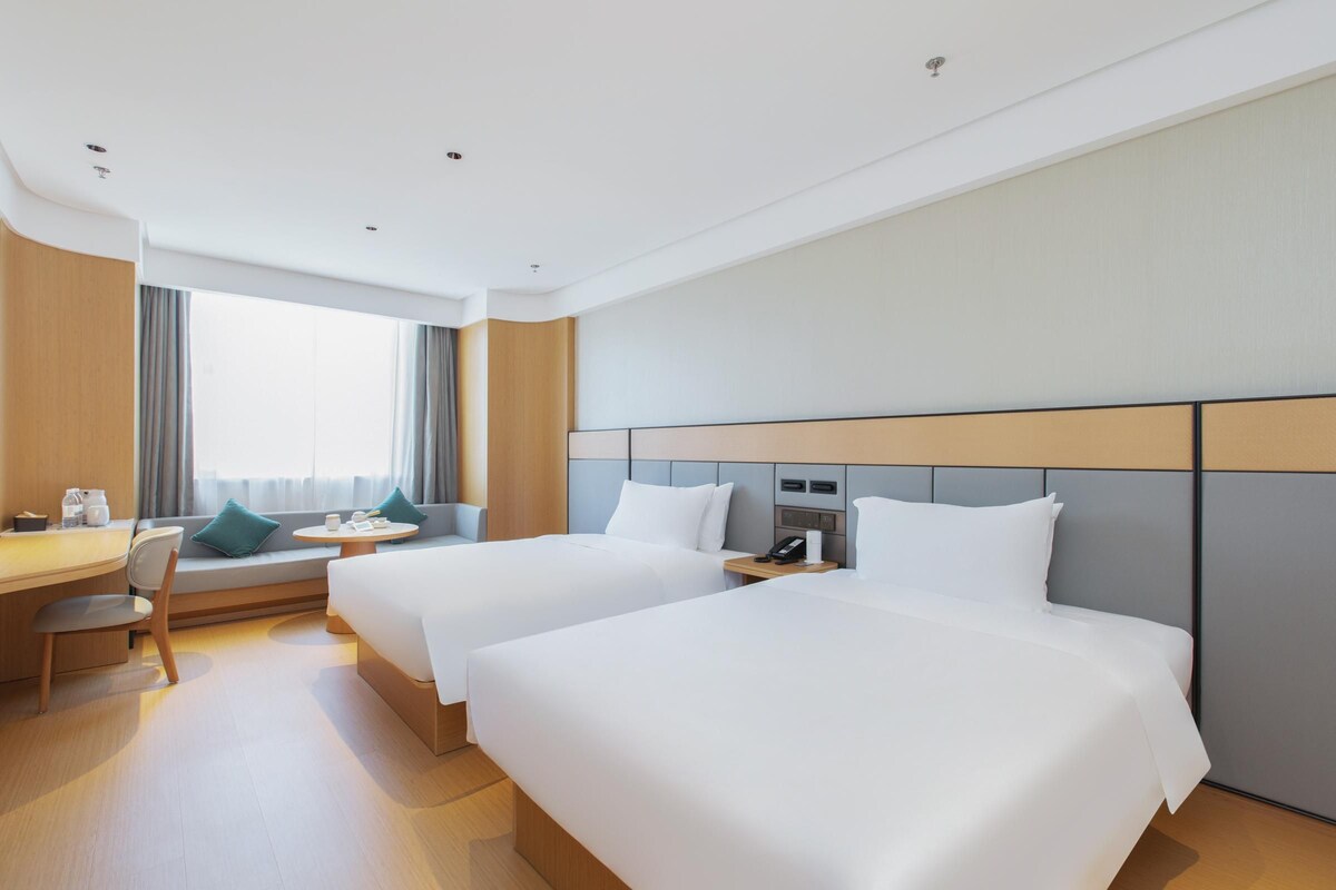 A hotel room with two beds, a desk, a chair, a small table, and a window with curtains.