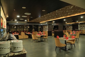 A restaurant with multiple tables and chairs, a buffet area with stacked plates, and a patterned ceiling. - Bravia Hotel Panchsheel (Ajmer)