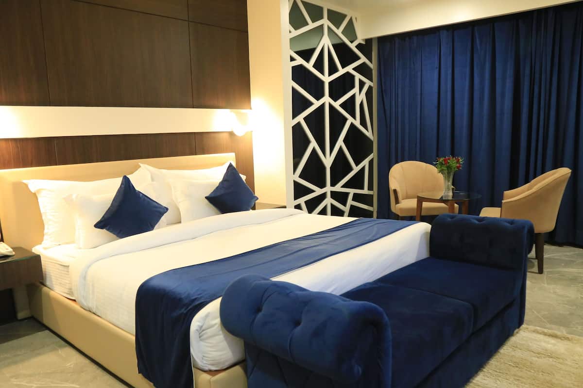 Habitación de hotel con una cama grande, una mesita de noche, una silla, una pequeña mesa con un jarrón y un diseño geométrico en la pared.