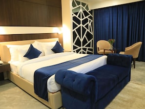 A hotel room with a large bed, a nightstand, a chair, a small table with a vase, and a geometric wall design. - Bravia Hotel Panchsheel (Ajmer)