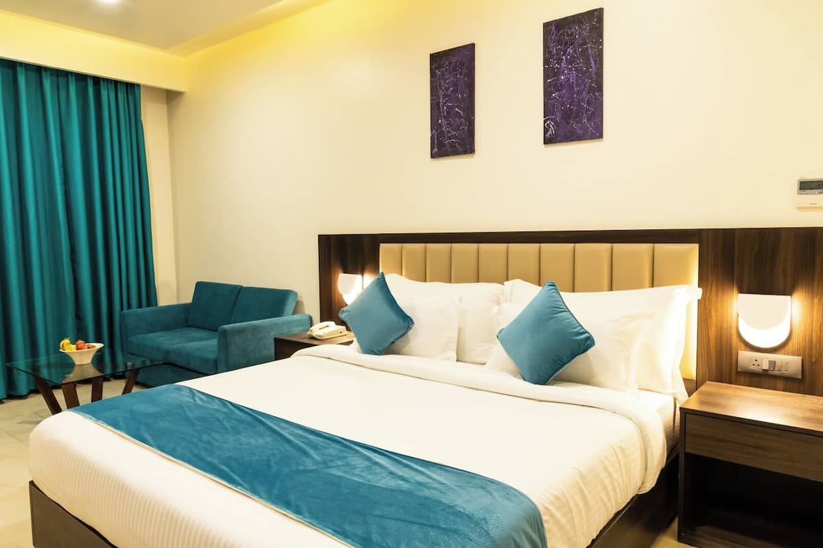 Habitación de hotel con cama, sofá azul, mesita de noche, lámpara y dos obras de arte en la pared.