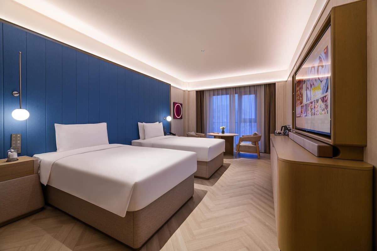 Une chambre d’hôtel avec deux lits, un bureau, une chaise, une télévision et une grande fenêtre avec des rideaux.