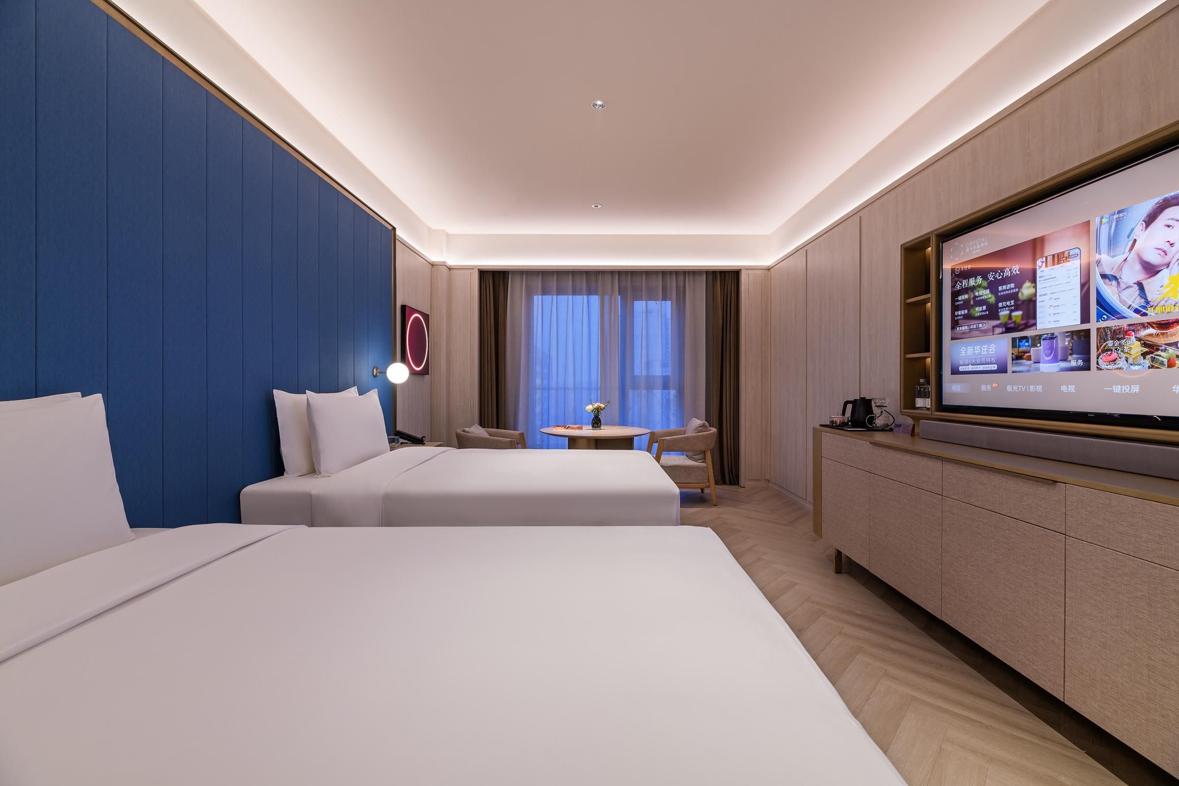 A hotel room with two beds, a TV, a small table with chairs, and a large window with curtains.