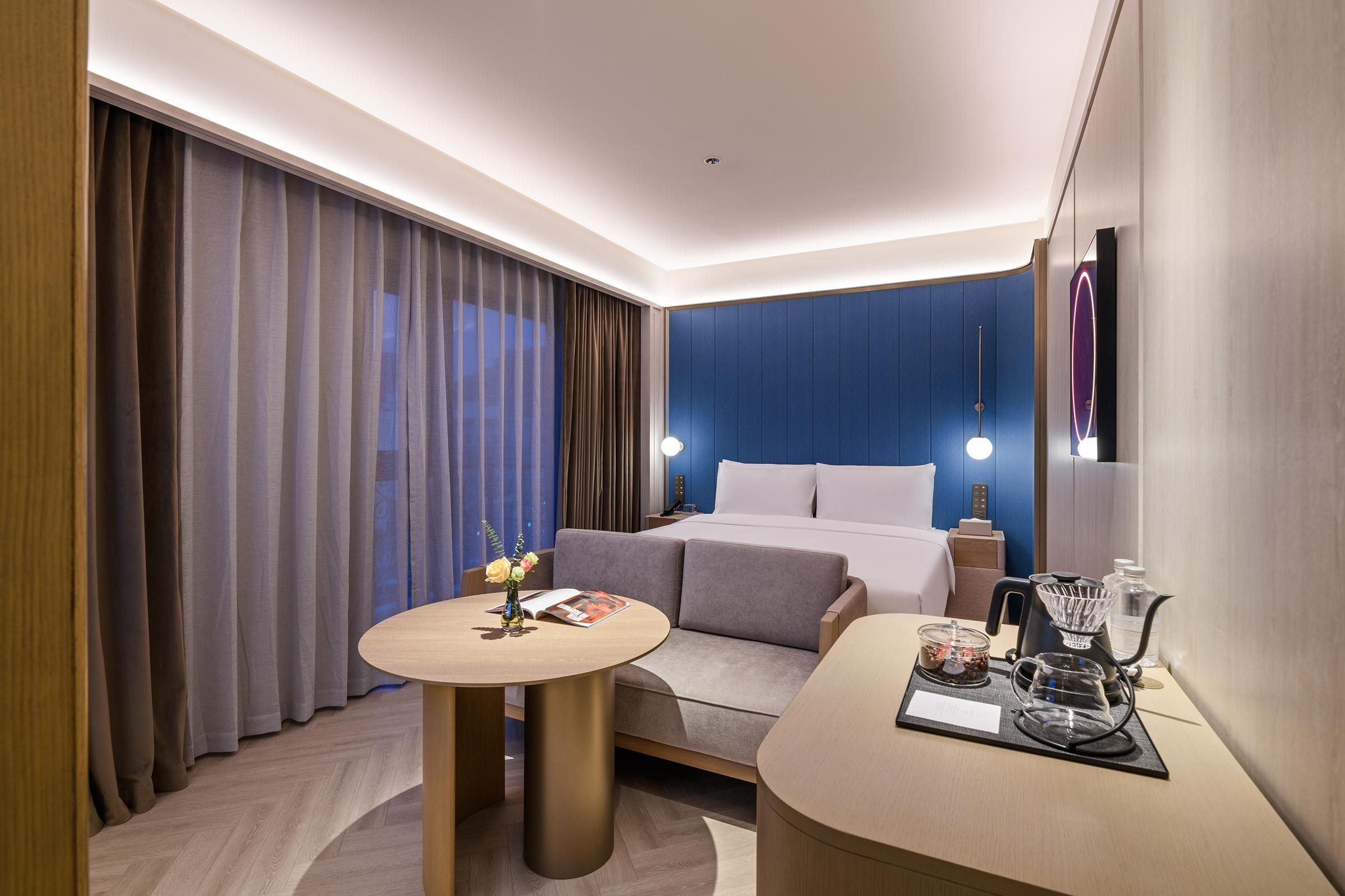 Een moderne hotelkamer met een bed, een klein tafeltje met een bloevas, een nachtkastje met een lamp en een koffiezetapparaat.