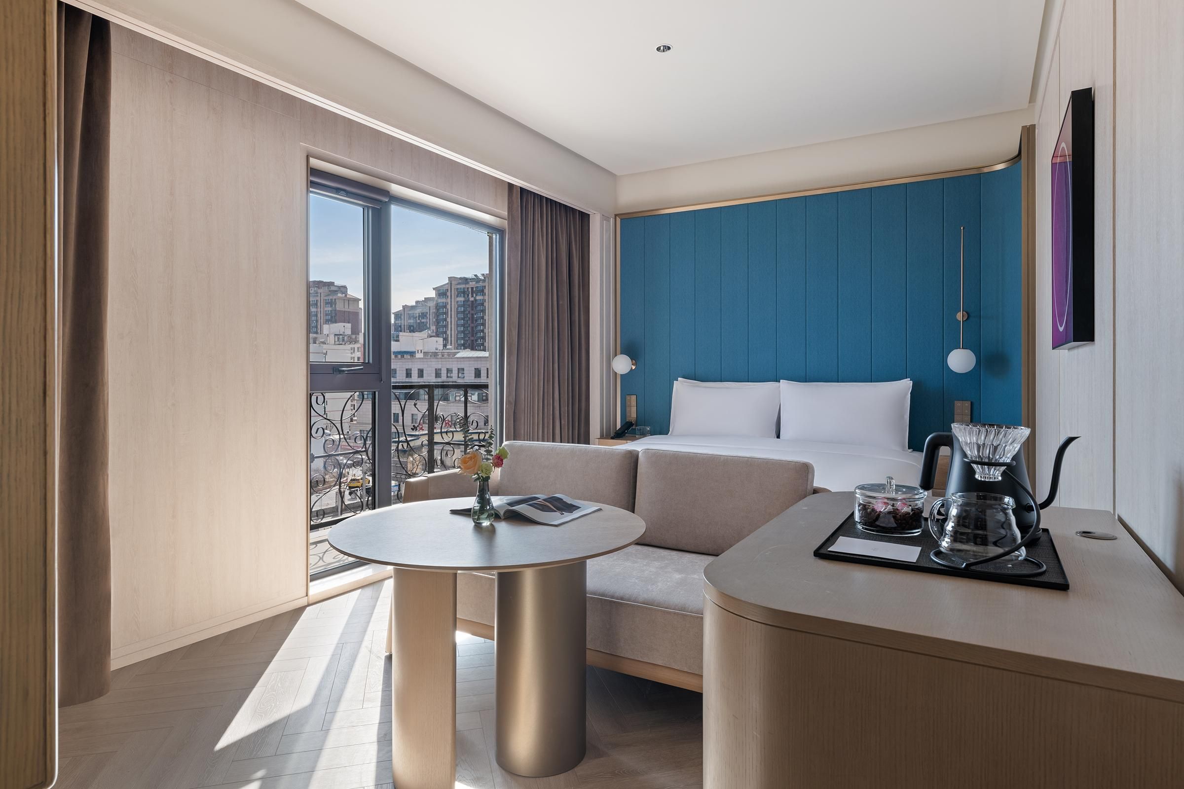 A modern hotel room with a large window, a sofa, a round table, and a coffee station.