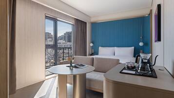 A modern hotel room with a large window, a sofa, a round table, and a coffee station.