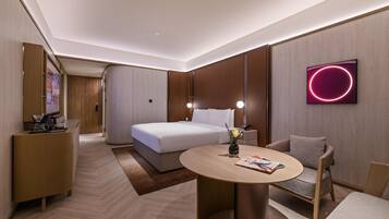 A modern hotel room with a bed, bedside tables, a round table, a sofa, and a wall-mounted TV.