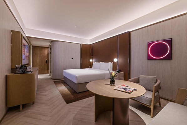 A modern hotel room with a bed, bedside tables, a round table, a sofa, and a wall-mounted TV.