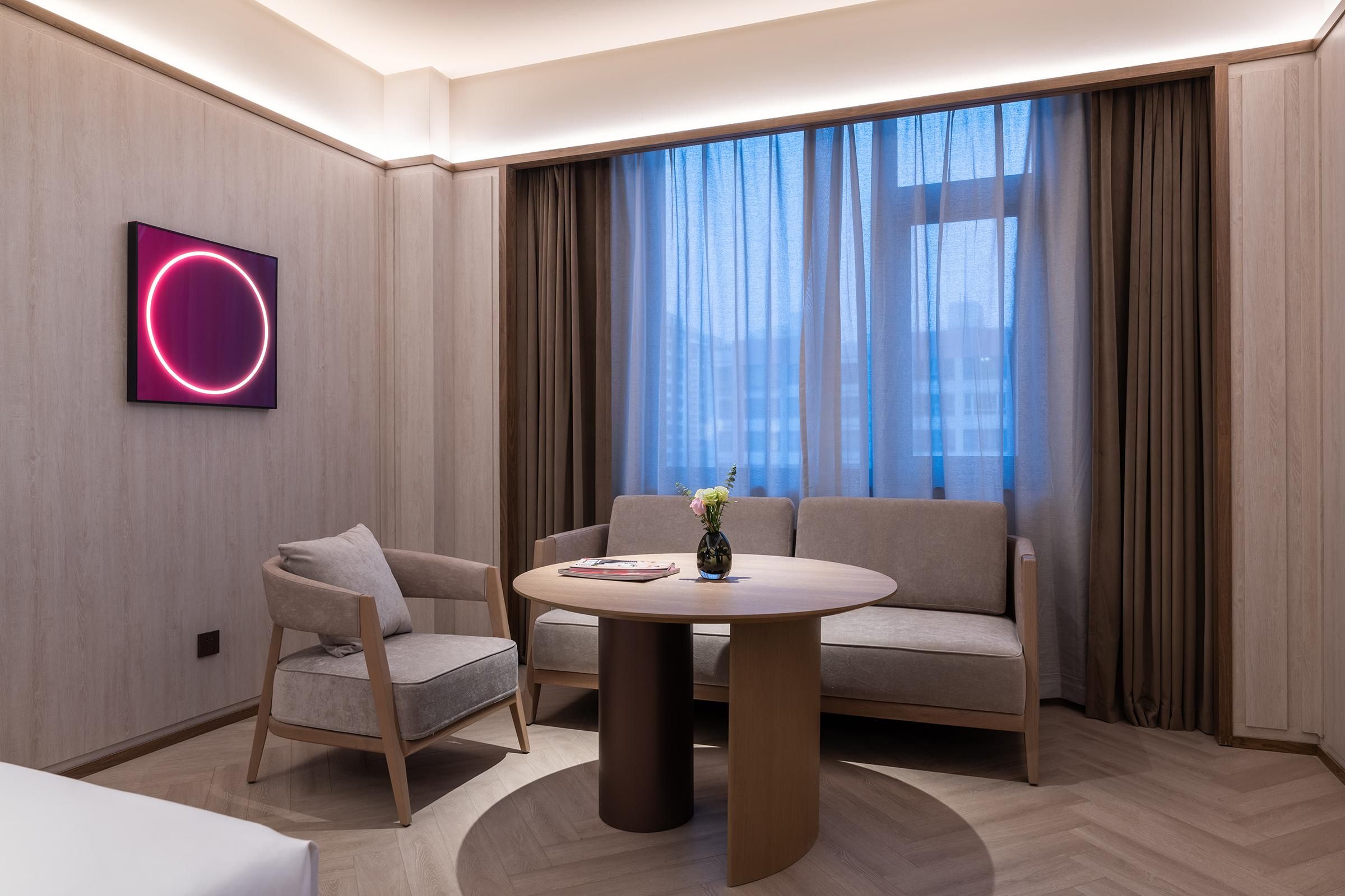 A modern hotel room with a round table, two sofas, a chair, and a large window with curtains.