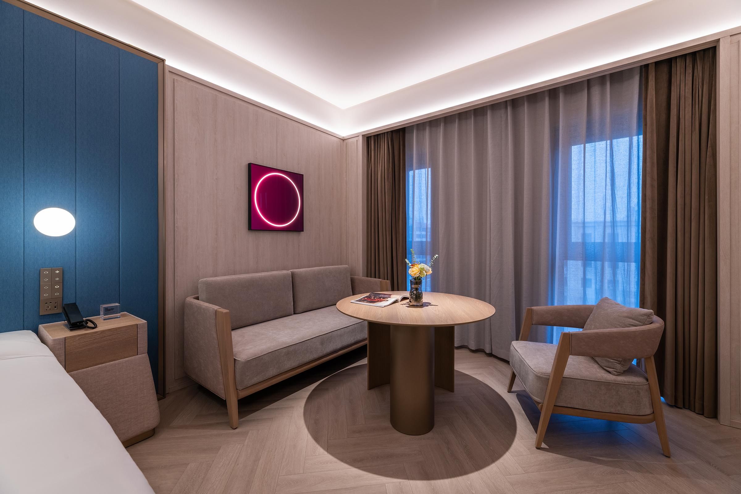 A modern hotel room with a round wooden table, a sofa, a chair, a nightstand, and a wall-mounted phone.