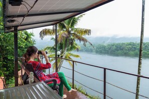 A person sitting on a chair by a railing overlooking a river, with a view of the surrounding greenery. - Munkudil Ayurveda Yoga Retreat (Kothamangalam)