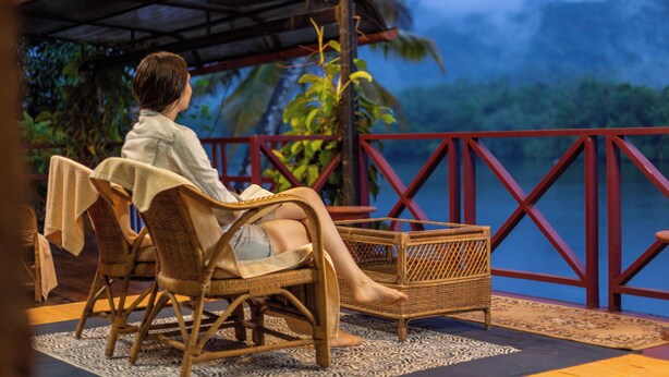 Una persona sentada en una silla de mimbre con una mesa, con vista a un cuerpo de agua y follaje.