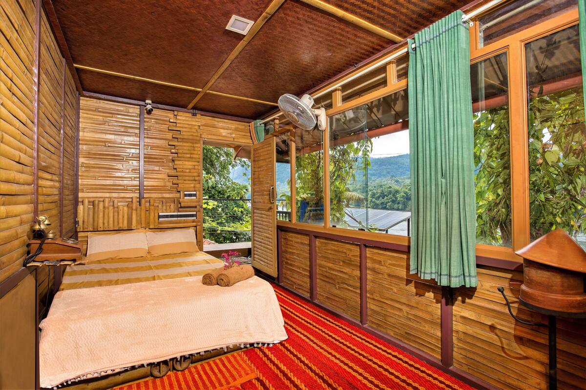 A wooden room with a bed, a window with a view, and a fan.