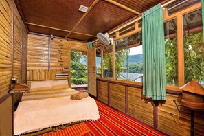 A wooden room with a bed, a window with a view, and a fan.