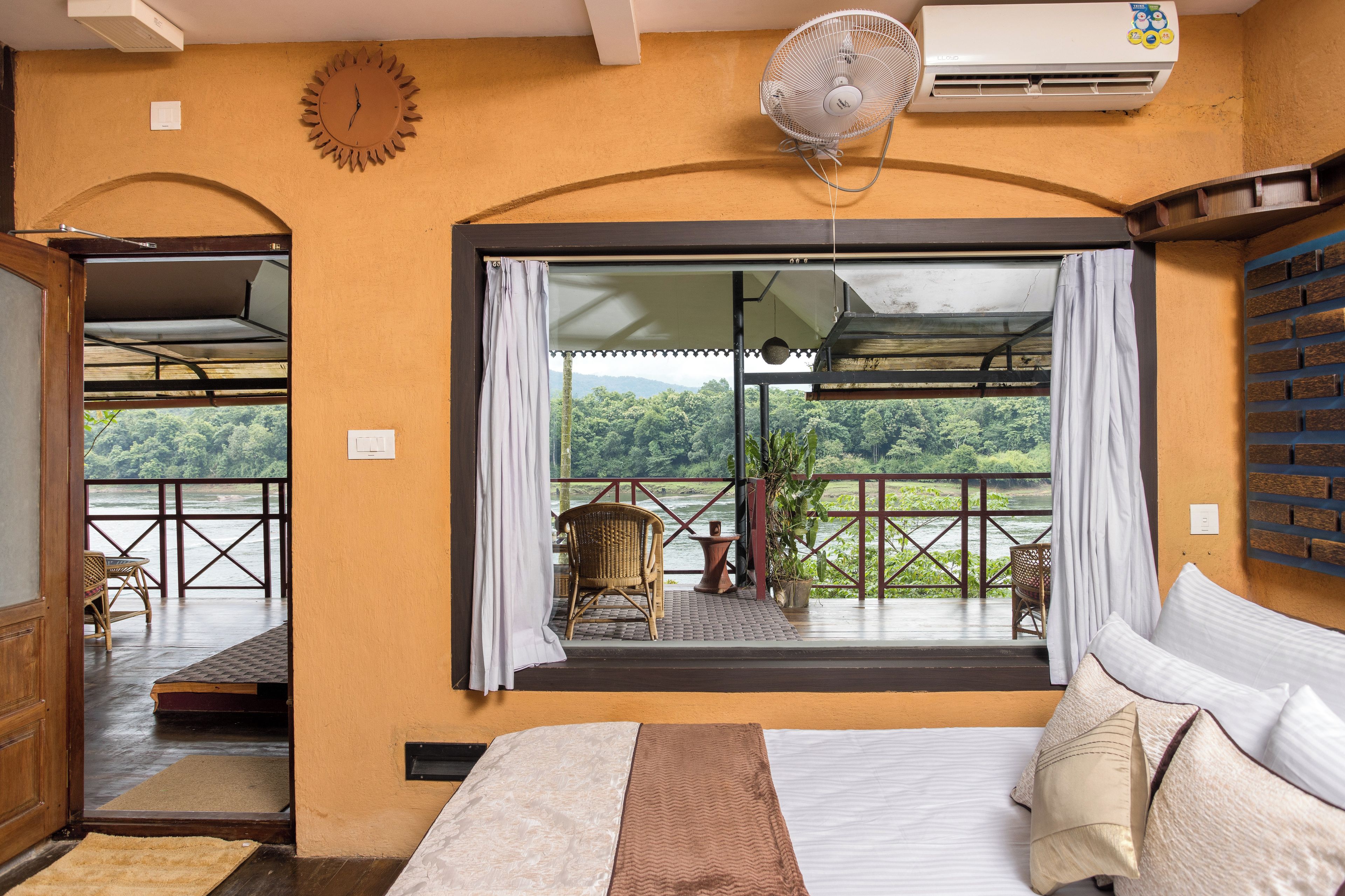 Quarto com cama, janela com cortinas, ventilador e vista para uma varanda com cadeiras e mesa.