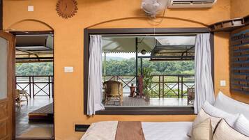 Quarto com cama, janela com cortinas, ventilador e vista para uma varanda com cadeiras e mesa.