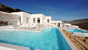 A modern white villa with a large infinity pool, outdoor seating, and a scenic mountain view. - Pili Pala Suites (Paros)