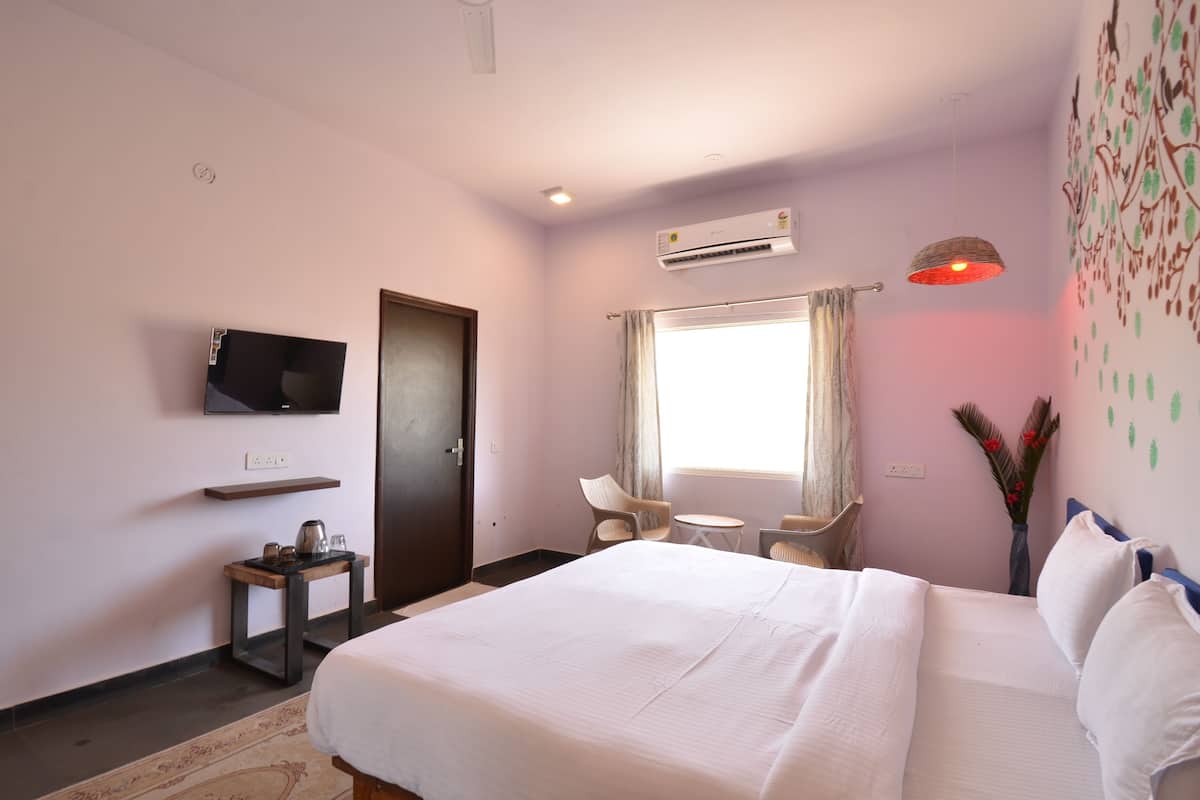 Habitación de hotel con cama, televisión, una mesita, silla, ventana con cortinas, aire acondicionado y un adorno en la pared.